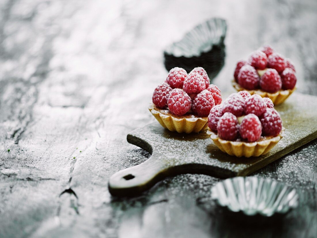 Three raspberry tartlets on a wooden board with tartlet tins