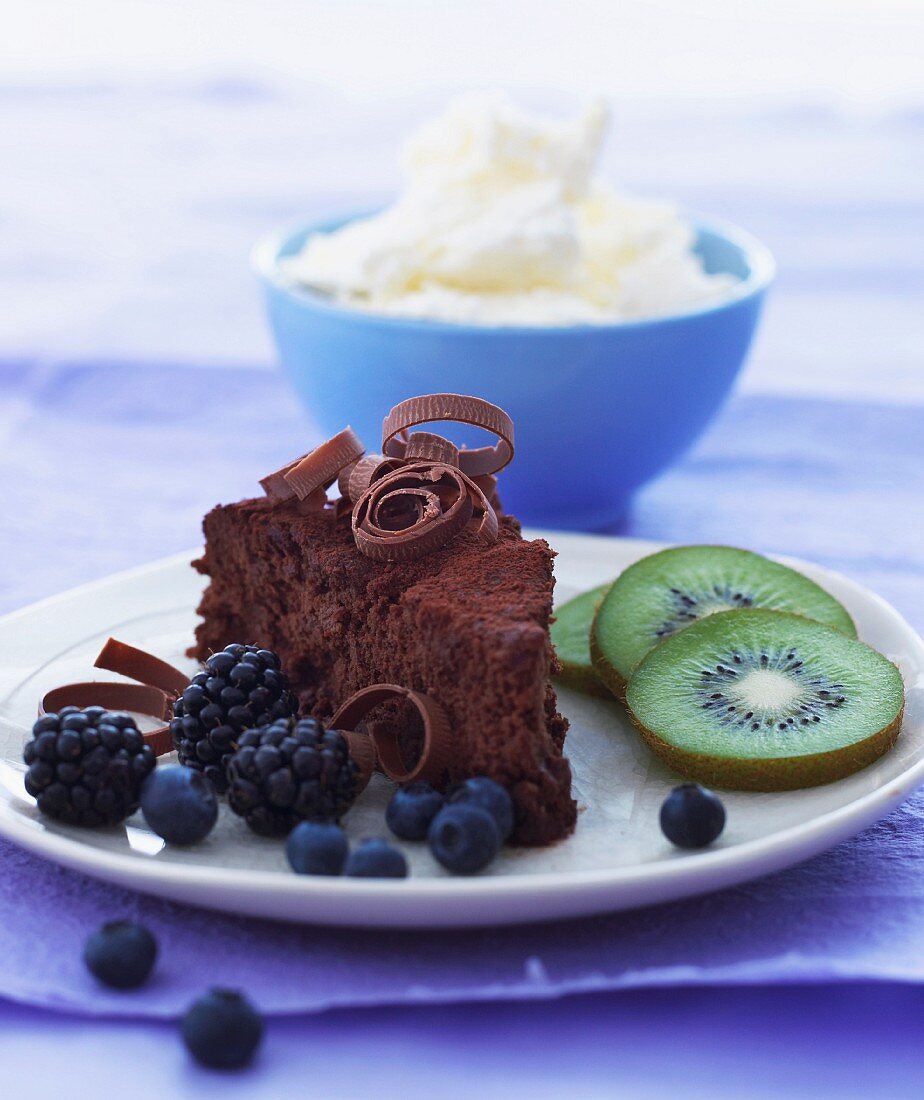 A slice of chocolate cake with fruit on a plate with a bowl of whipped cream in the background