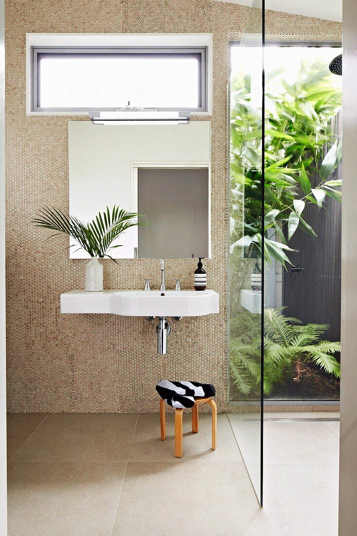 Sink on bathroom wall covered in round, beige iridescent mosaic tiles; walk-in shower with glass wall looking into planted courtyard