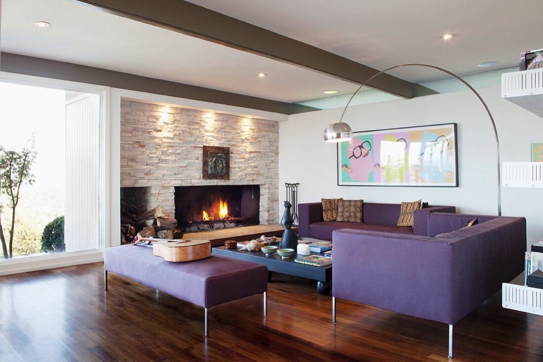 Elegant, purple sofa set in lounge area with arc lamp and cosy open fire in stone wall