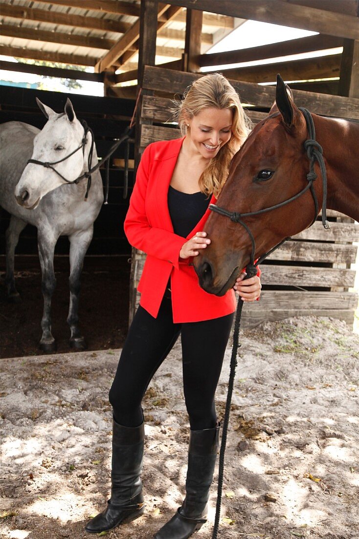Junge, blonde Frau mit roter Jacke neben Pferd vor dem Stall