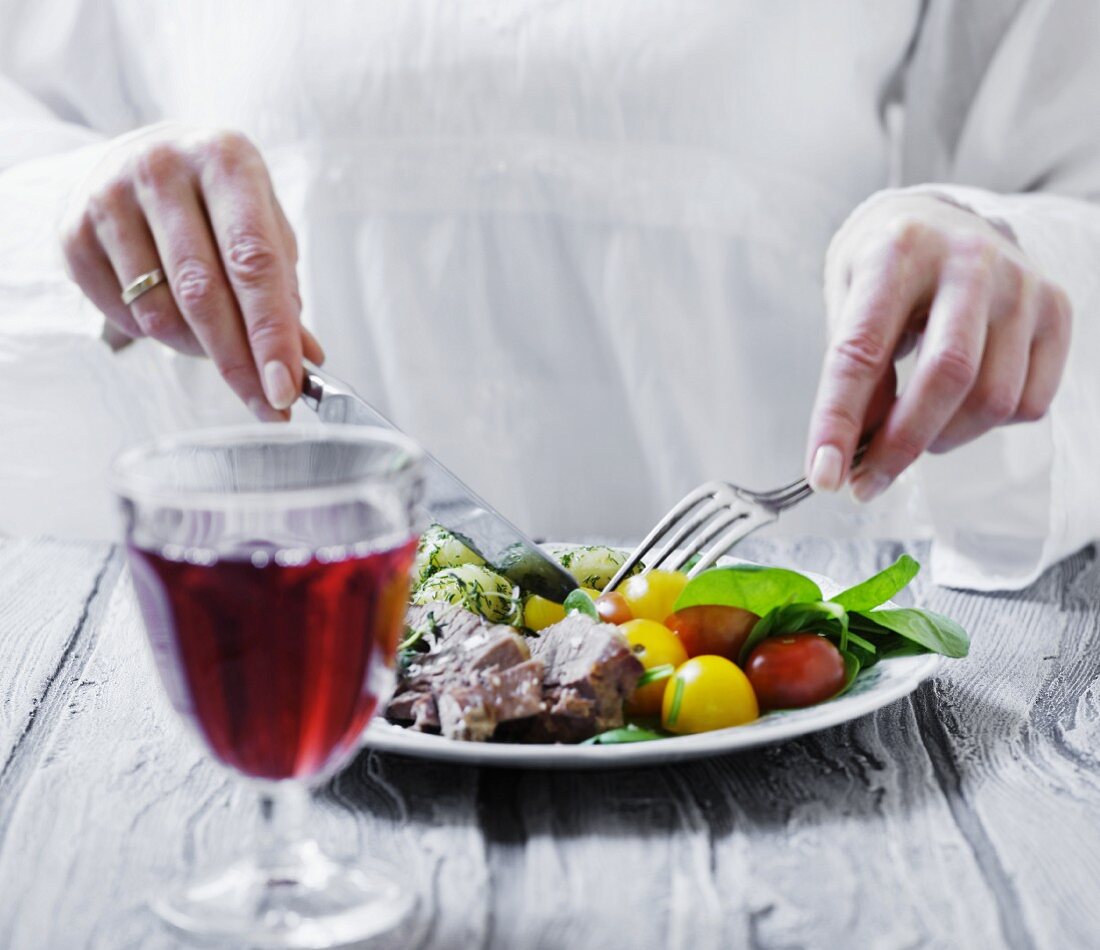 Glas mit Rotwein vor einem Teller mit Rindfleisch, Tomatensalat und Dillkartoffeln