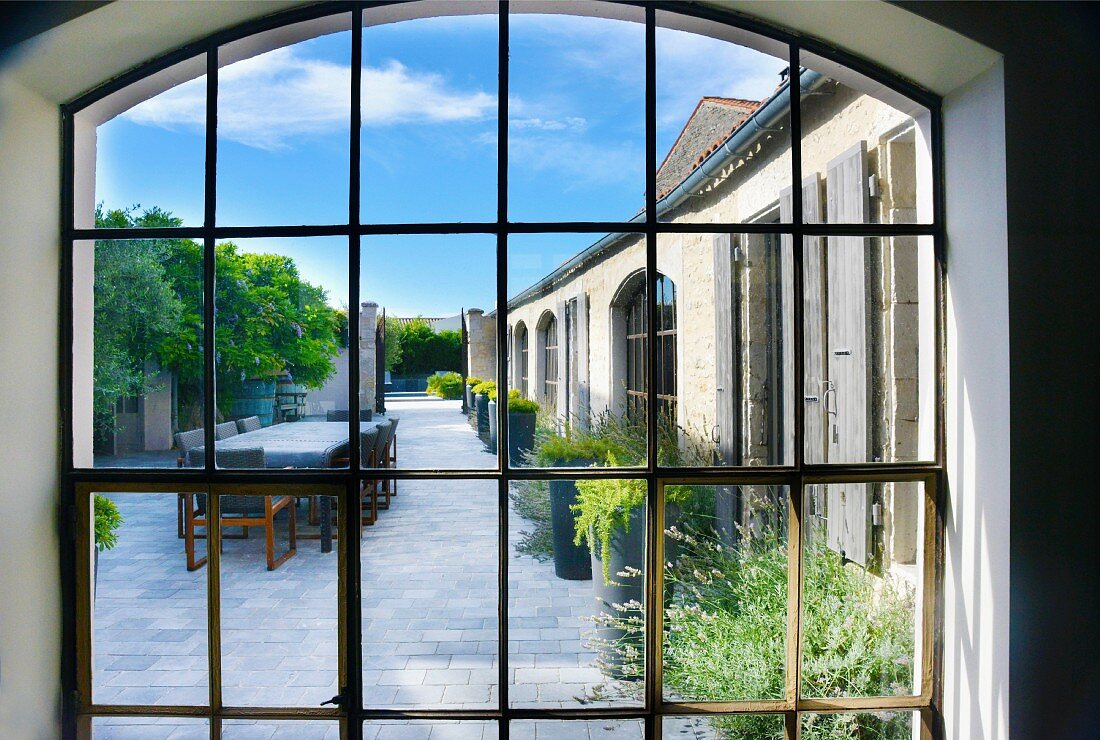 View through lattice window into courtyard with planters along bungalow facade and long table and chairs on flagged floor