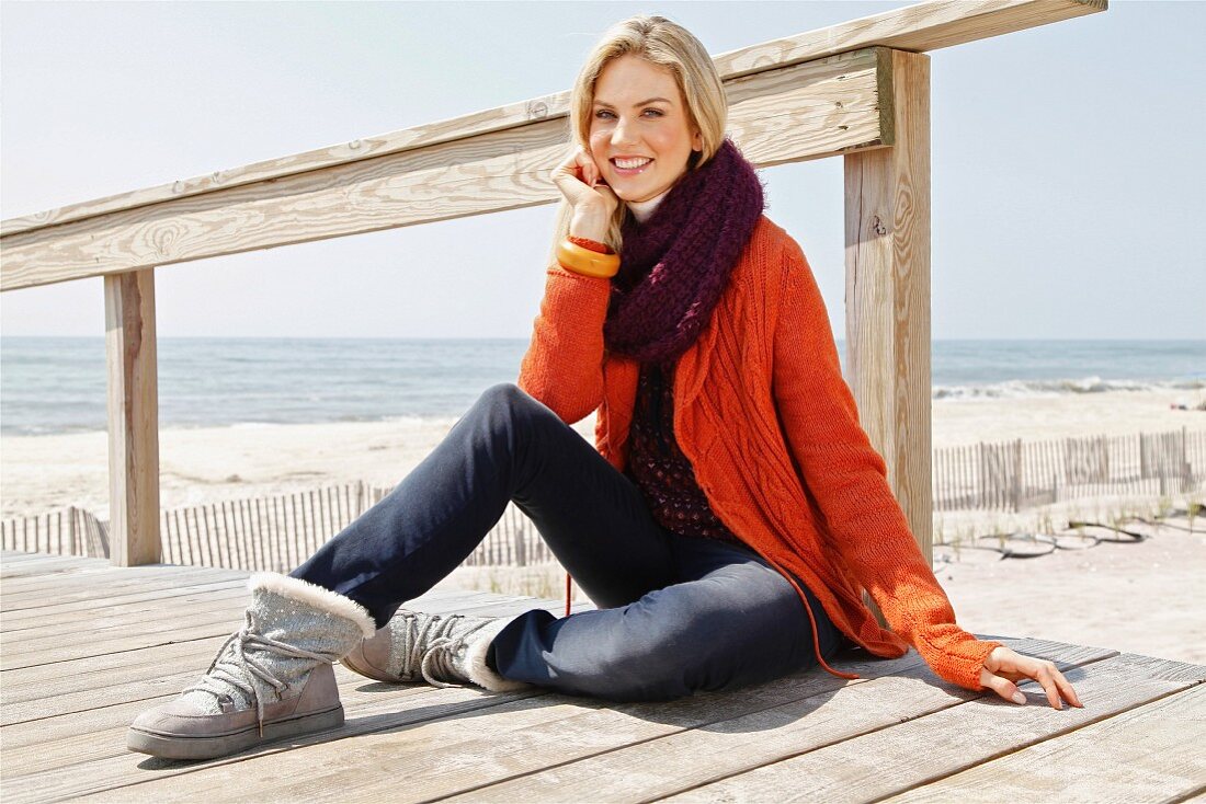 A young blonde woman sitting on a wooden boardwalk wearing an orange cardigan and a loop scarf