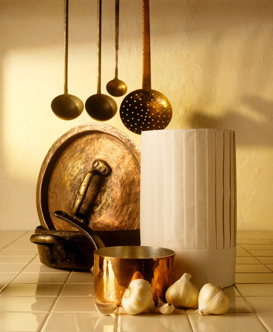 A chef's hat, copper pots, ladles and garlic