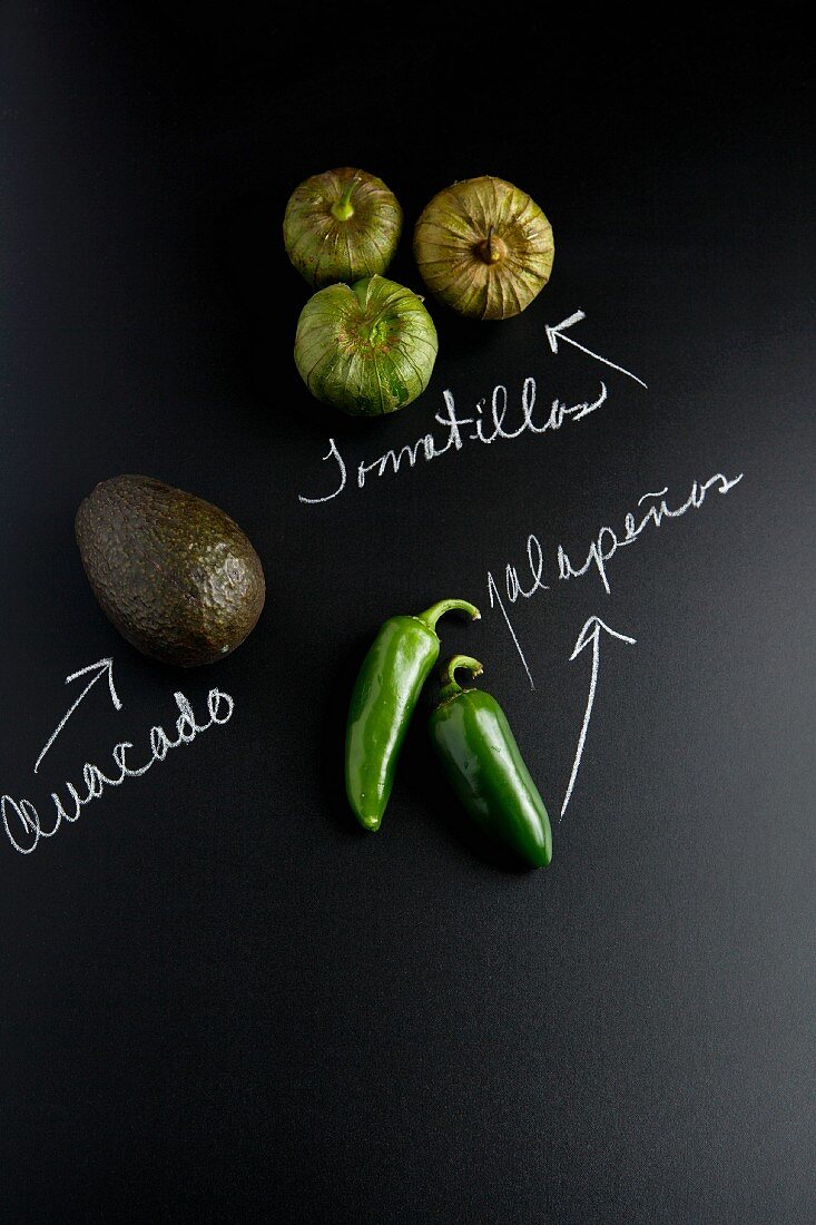 An avocado, jalapeños and tomatillos on a slate surface with labels