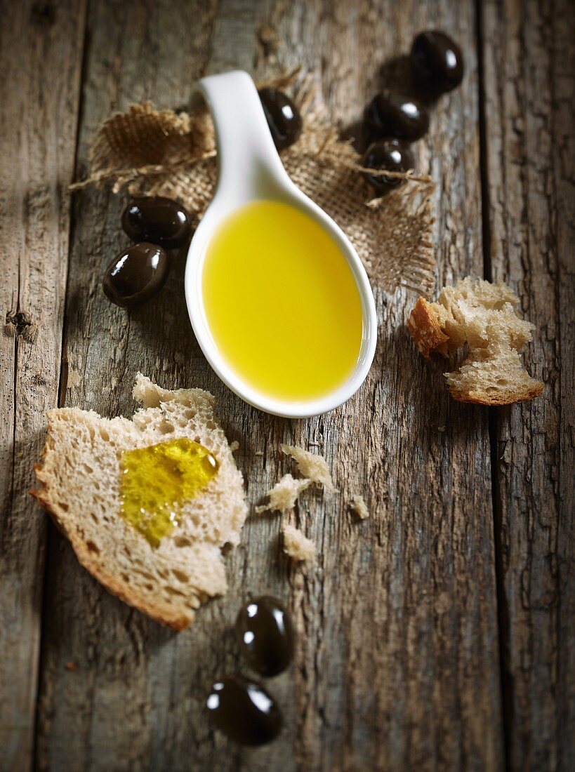 A spoonful of olive oil, bread and black olives on a wooden surface