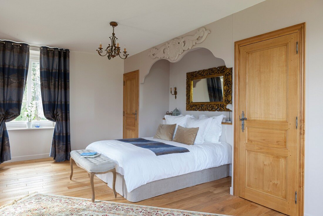 Elegantes Doppelbett in Nische mit Goldrahmenspiegel und geschnitztem Ornament, im Hintergrund bodenlanger blaugrauer Vorhang an Fenster