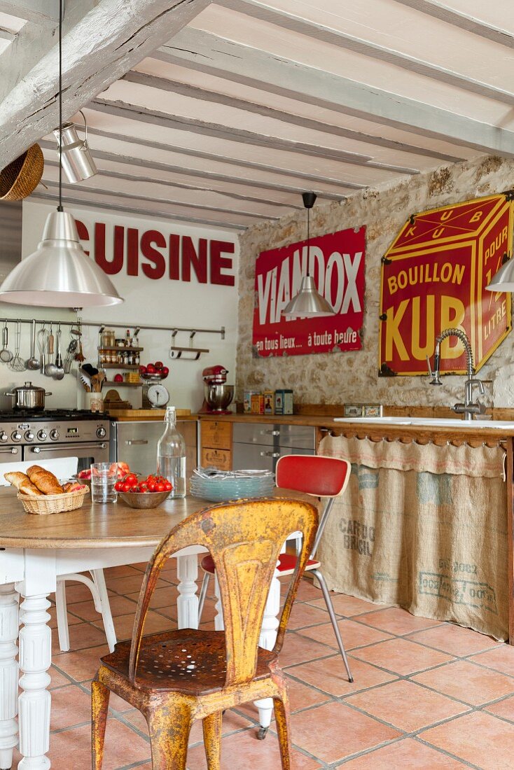 Chairs of various styles around rustic table, kitchen counter with curtains on base units and vintage advertising signs