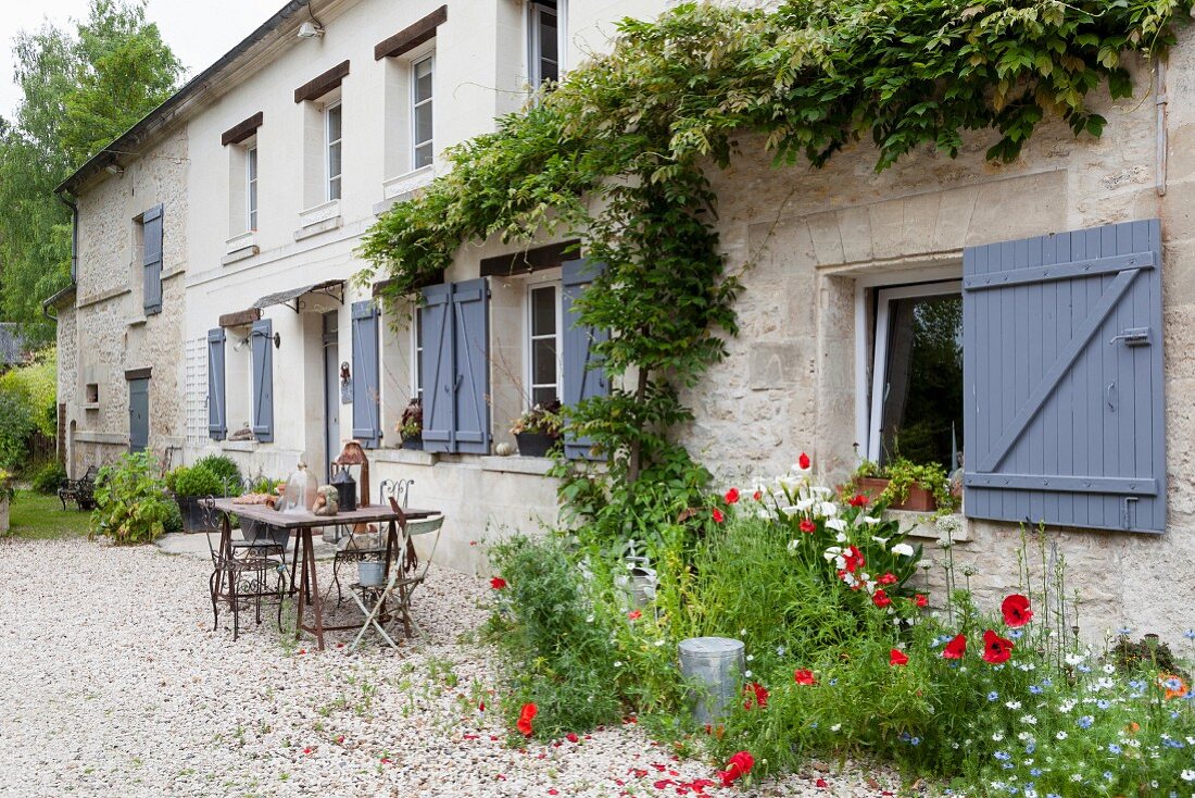 Poppies outside rustic country house with window shutters painted slate grey and seating area on gravel terrace