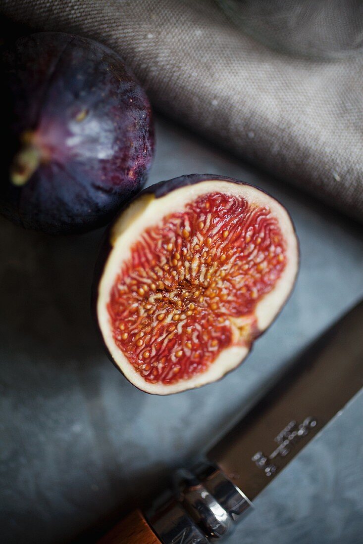 A whole fig and a halved fig on a galvanised surface with a linen cloth and a knife