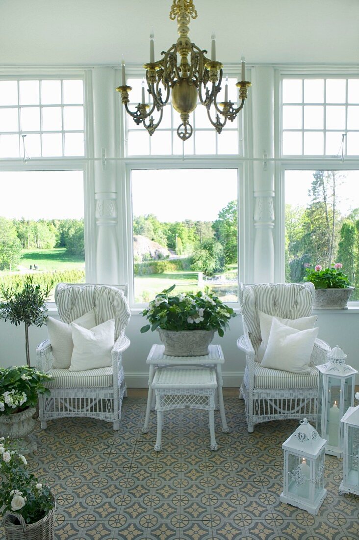 Chandelier above white, wicker chairs, set of matching tables and lanterns in conservatory with view into sunny park