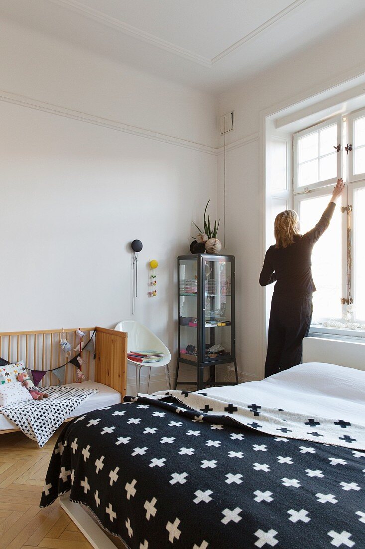 Bedspread with pattern of crosses and cot in bedroom; woman opening window in background