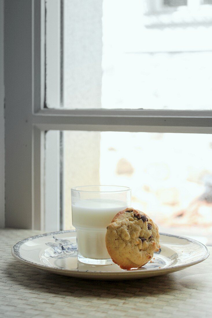 A chocolate chip cookie and a glass of milk in front of a window