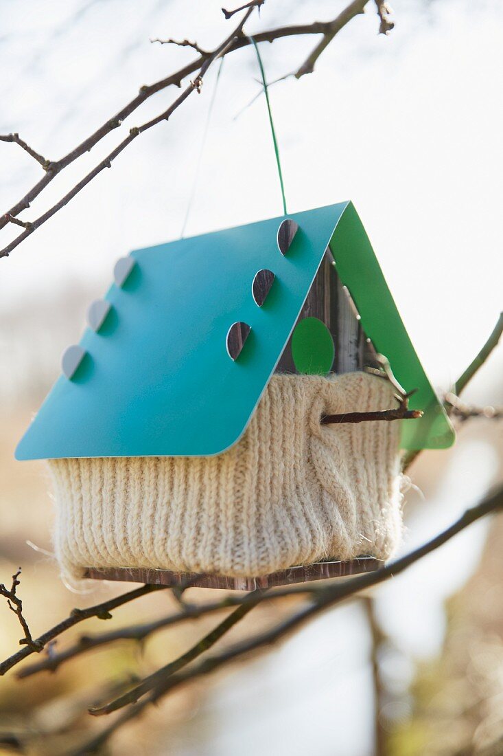 A birds house with a knitted cover hanging on a twig