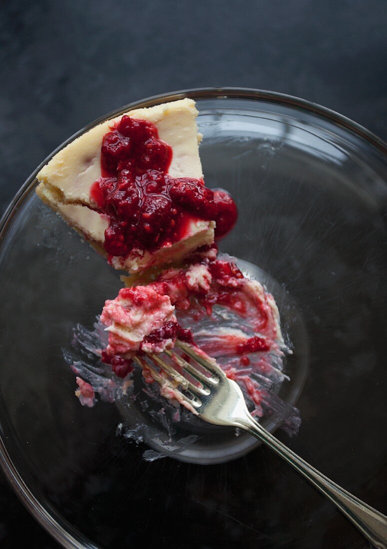 A slice of cheesecake with raspberry sauce on a glass plate with a fork with a bite taken out