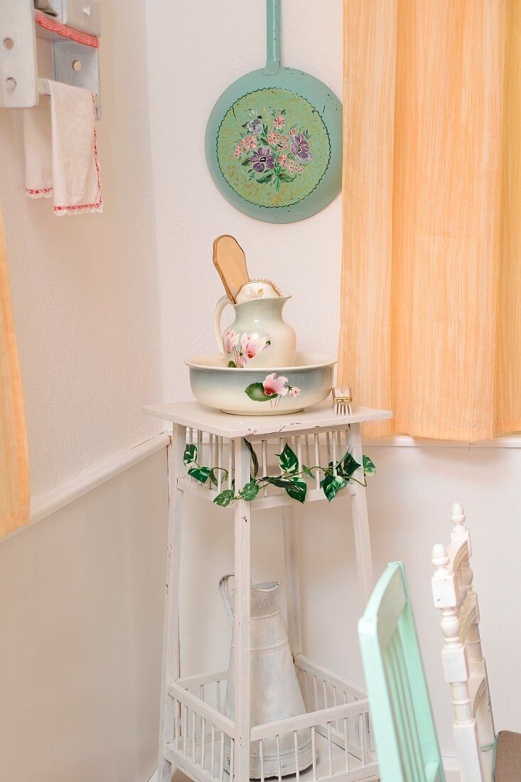 Old china pitcher and basin set and large enamel jug on plant stand below painted frying pan on wall in corner of shabby-chic kitchen