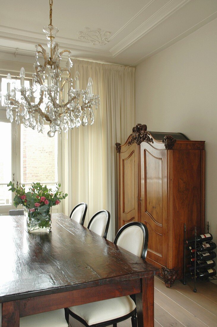 Rustic wooden table and neo-antique, upholstered chairs below crystal chandelier in grand dining room