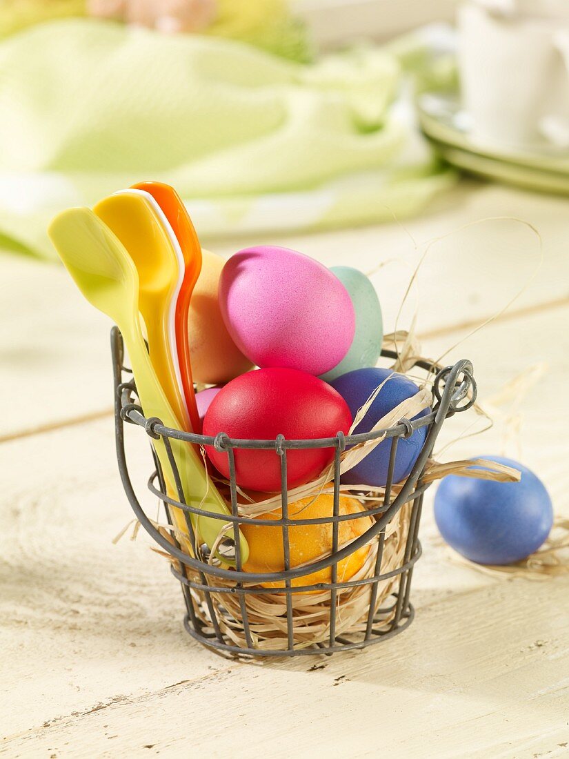 Colourful Easter eggs and plastic spoons in a wire basket lined with straw