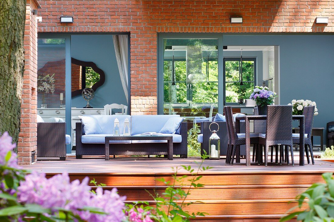 Moderne Outdoormöbel aus dunklem Rattan auf Holzterrasse, vor Wohnhaus mit grau lackierten Fenstern und Wandflächen in Ziegelfassade