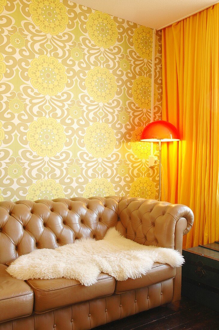 White, sheepskin blanket on brown, leather, button-tufted sofa; standard lamp with orange lampshade in corner against 70s-style, patterned wallpaper