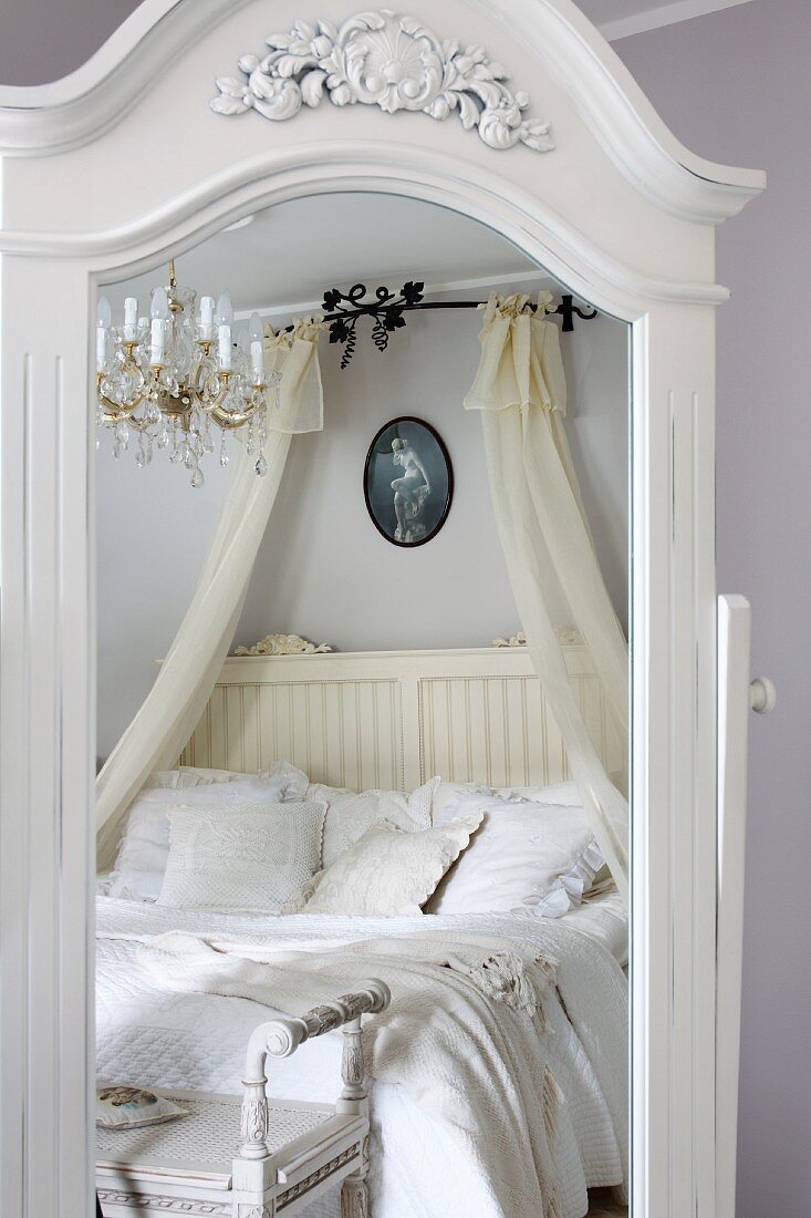 Spiegel mit weißem, geschnitztem Holzrahmen, darin reflektiertes Doppelbett mit weisser Bettwäsche und Baldachin