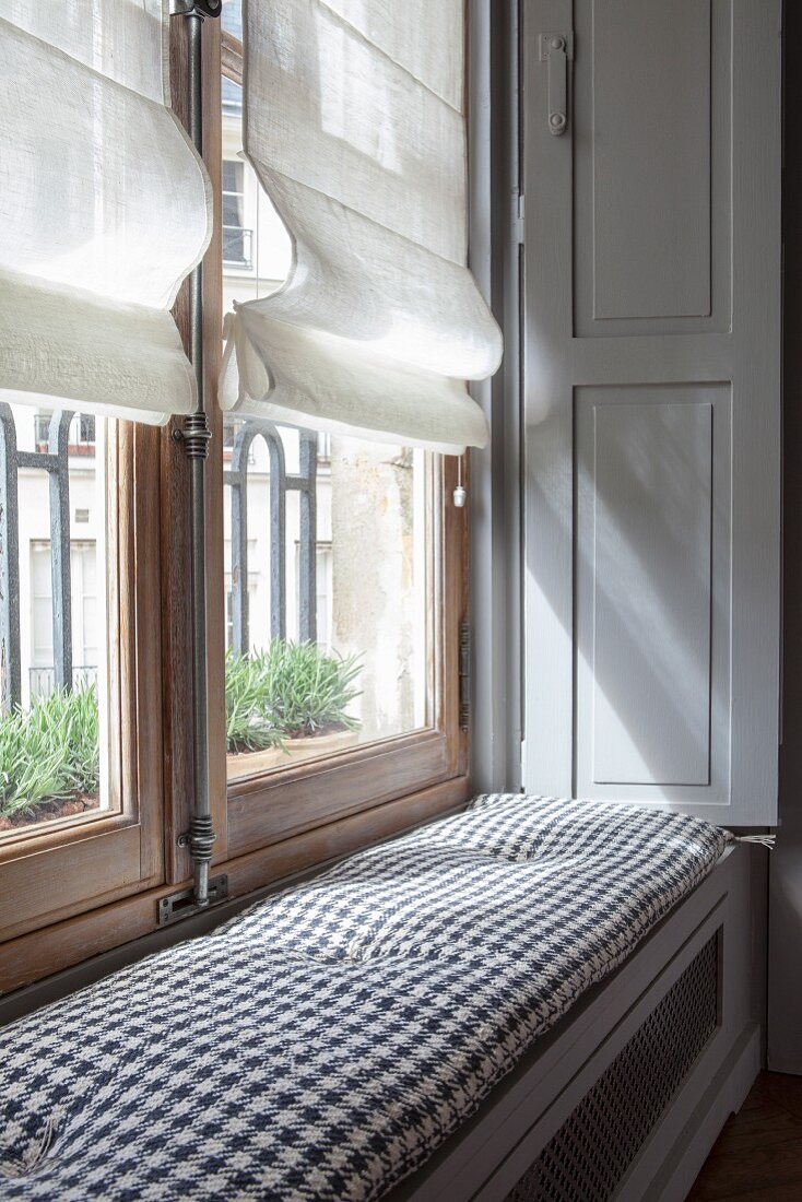 Window seat with cushion on top of radiator cover in niche of window in period apartment