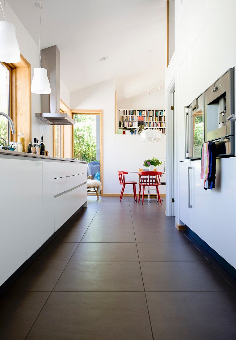 Blick mit Untersicht in moderner Küche, zwischen Küchenzeile und eingebauten Küchengeräten, im Hintergrund Essplatz mit roten Stühlen