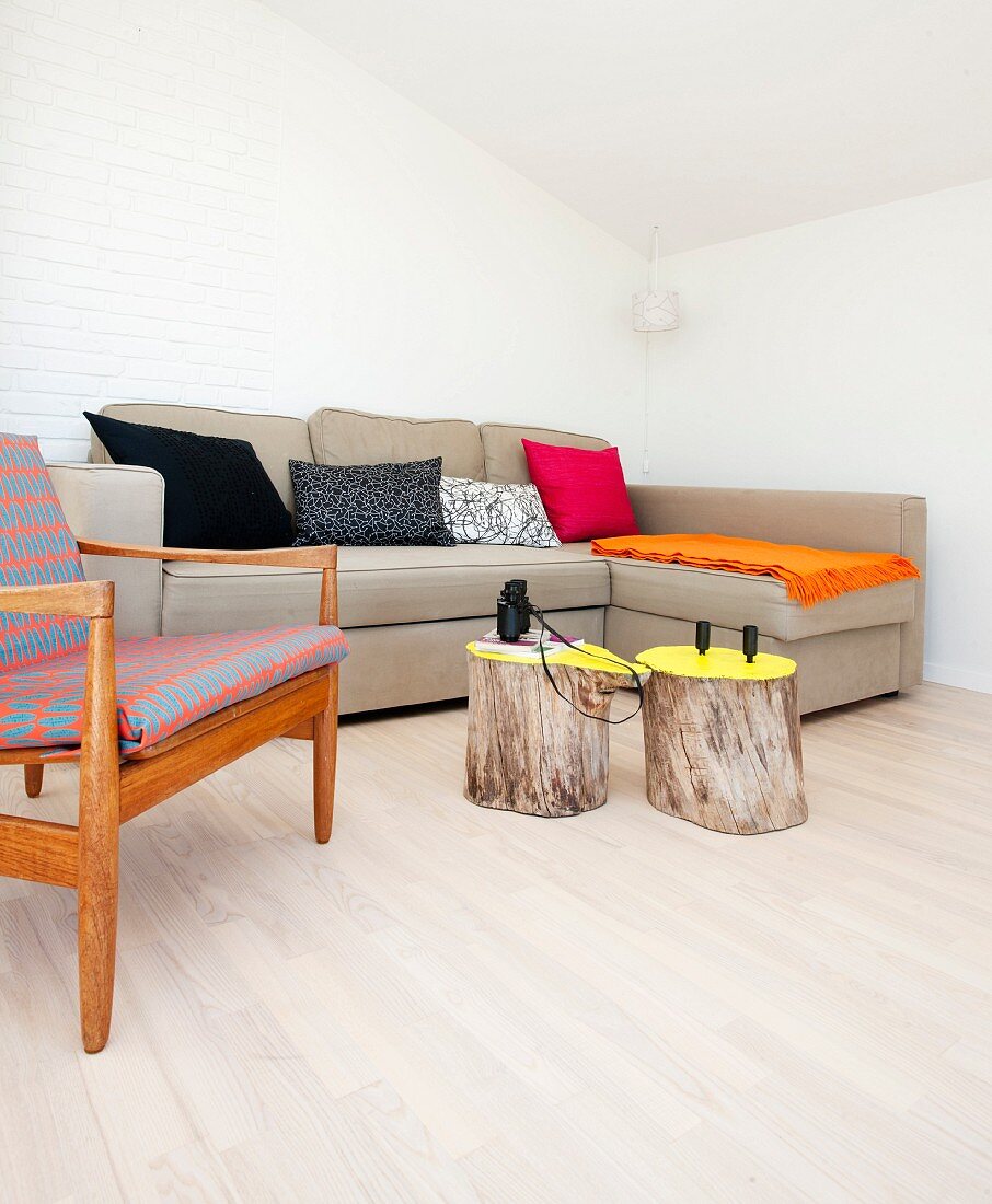Baumstämme als Beistelltisch vor naturfarbenem Sofa in minimalistischer Wohnzimmerecke, seitlich Sessel im Fiftiesstil