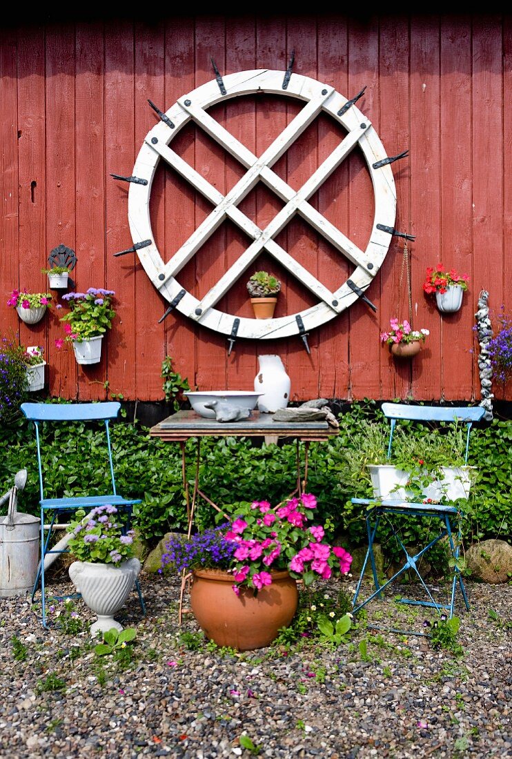 Old mill wheel and potted plants on Falu-red wooden wall behind garden furniture and flowers
