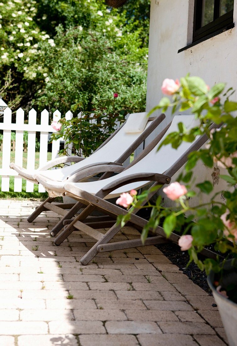 Wooden deckchairs with white seats on terrace outside house with garden in background