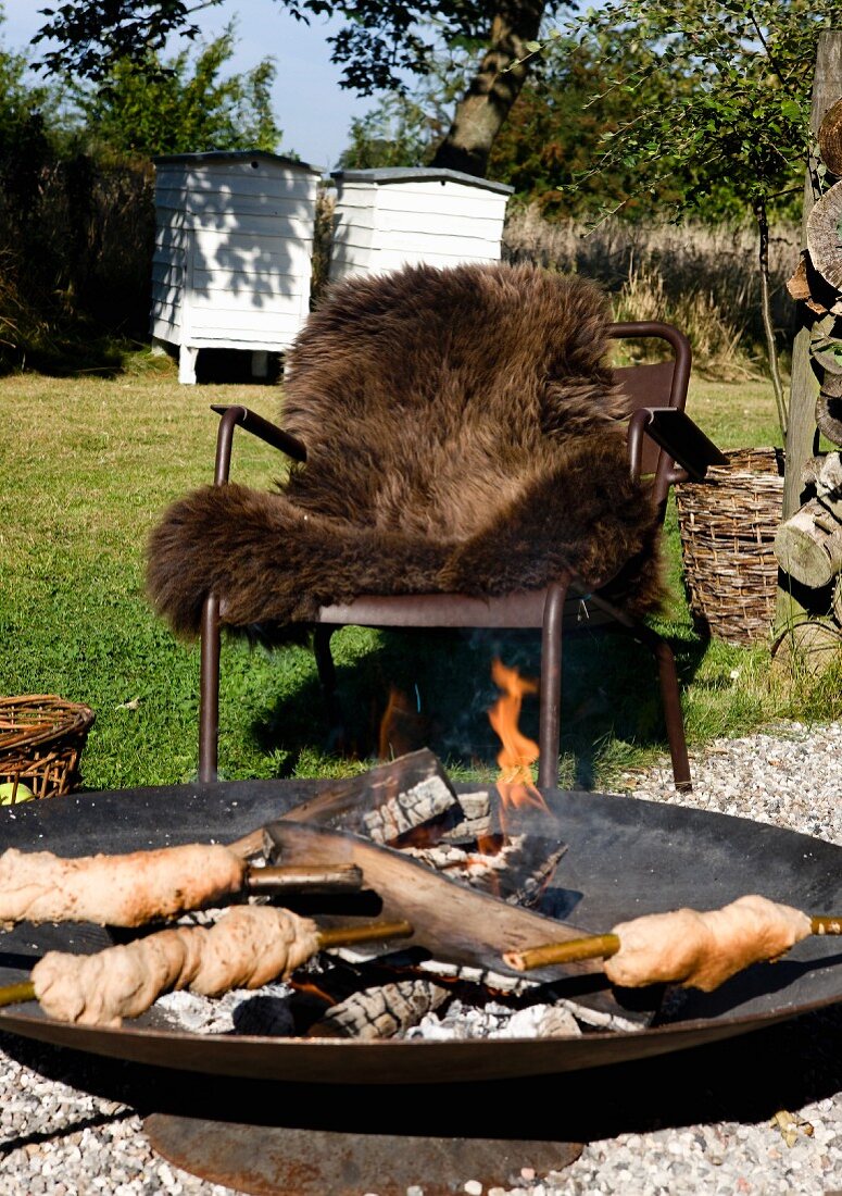 Stockbrot in Feuerschale, dahinter Outdoor Sessel mit braunem Tierfell im Garten