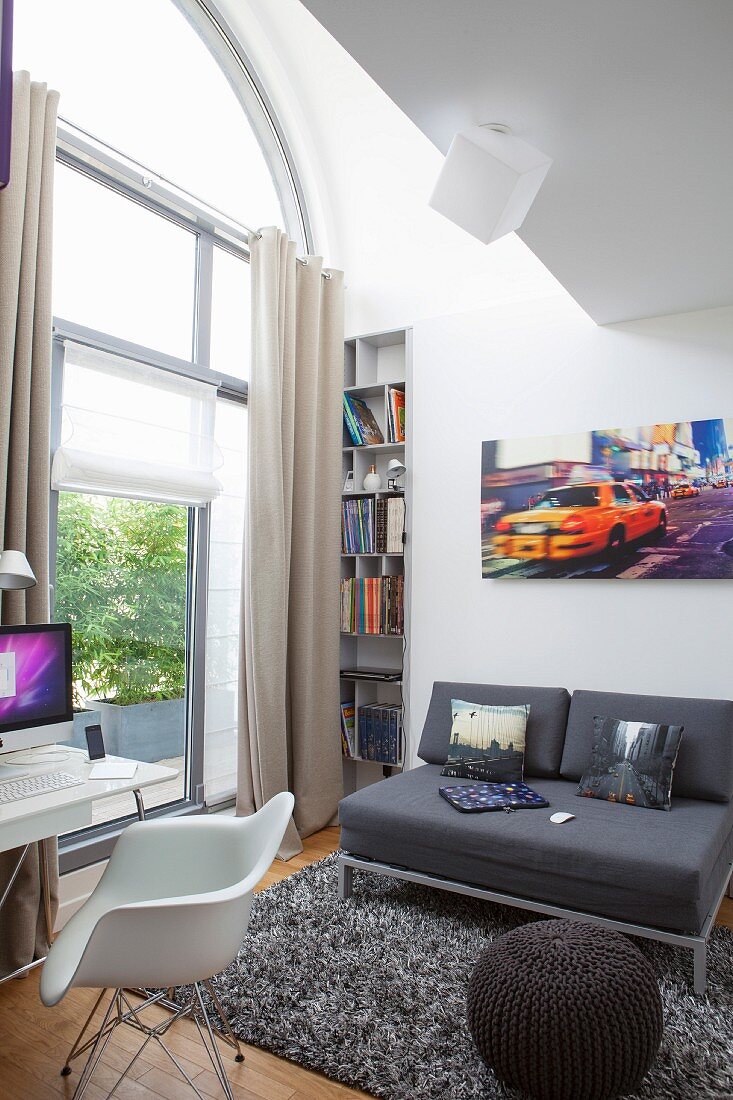 Sofa and knitted pouffe on long-pile rug next to tall, arched window with shell chair and desk in foreground