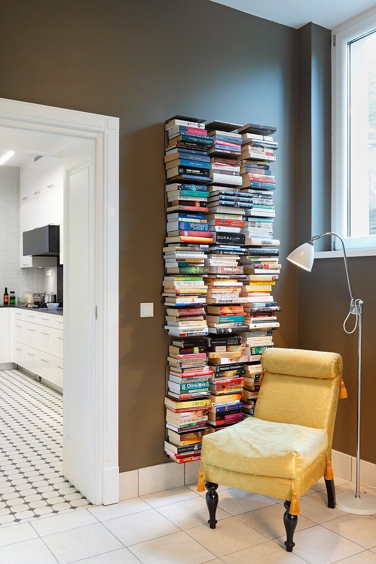 Antique upholstered easy chair and modern standard lamp in front of books stacked on narrow wall-mounted shelves; view into modern kitchen to one side
