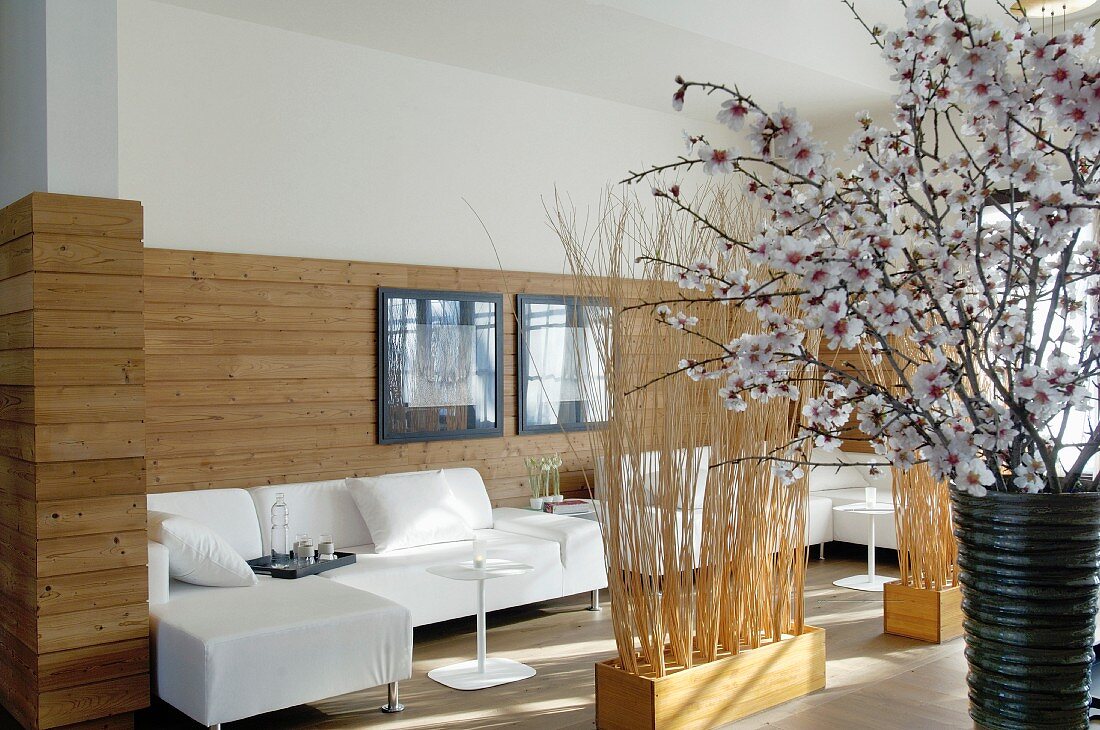 Half-height wood cladding behind white sofa combination, partitions made of bundled brushwood and vase of cherry blossom in foreground