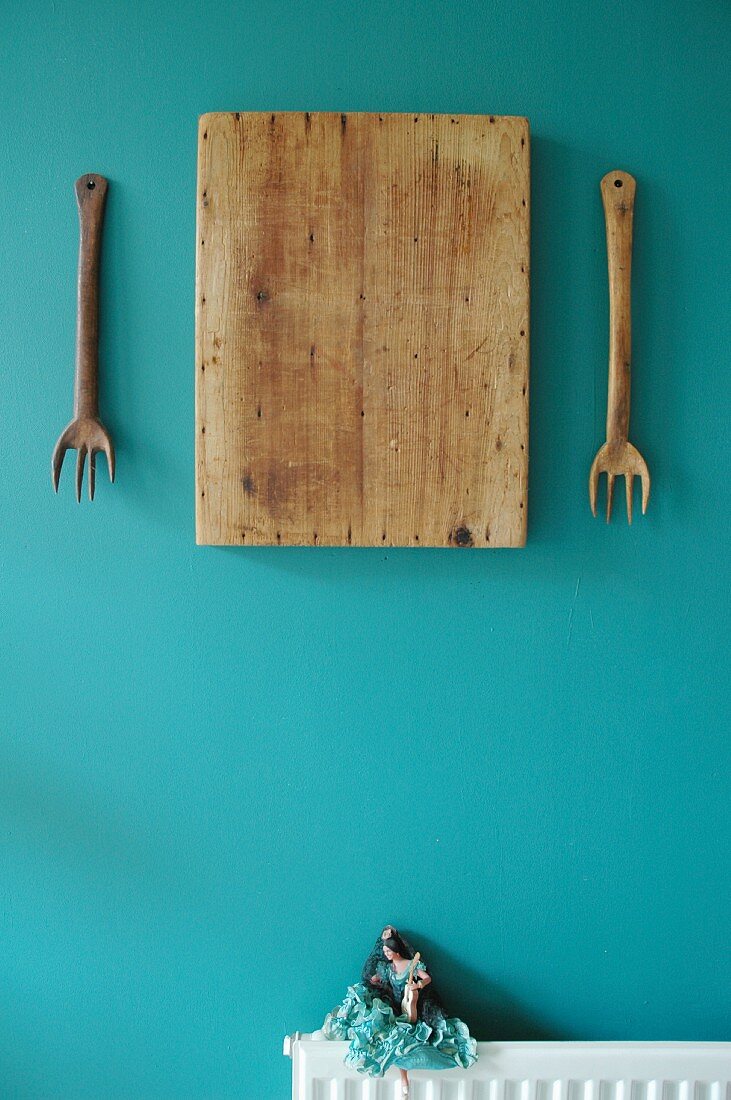 Artistic still-life arrangement of wooden forks and old chopping board on turquoise wall above tiny Flamenco doll sitting on radiator
