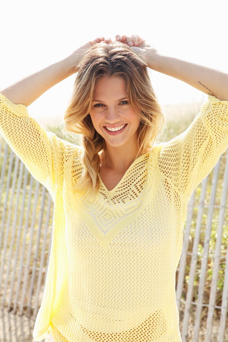 A young blonde woman on a beach wearing a white top and a pastel-yellow openwork jumper
