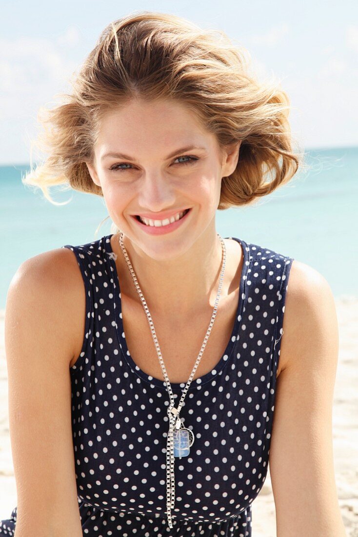 A young blonde woman on a beach wearing a sleeveless, polka-dot summer dress