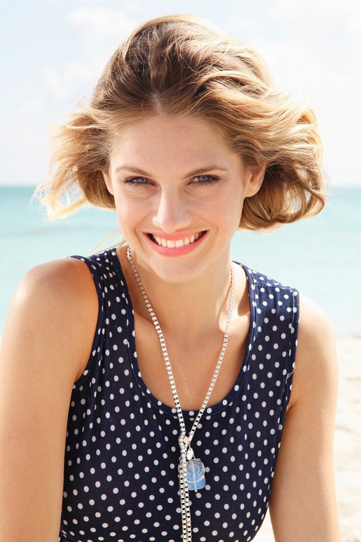 Junge blonde Frau in ärmellosem Sommerkleid mit Tupfen am Strand