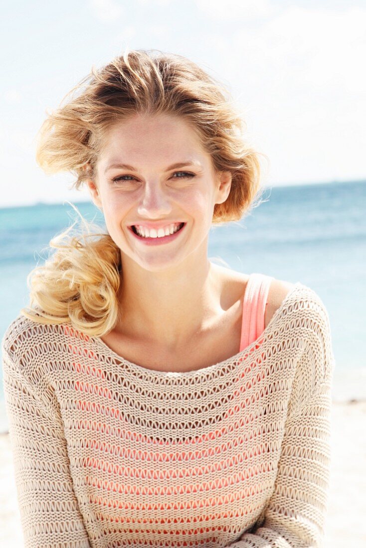 A young blonde woman on a beach wearing a salmon-coloured top and a beige openwork jumper