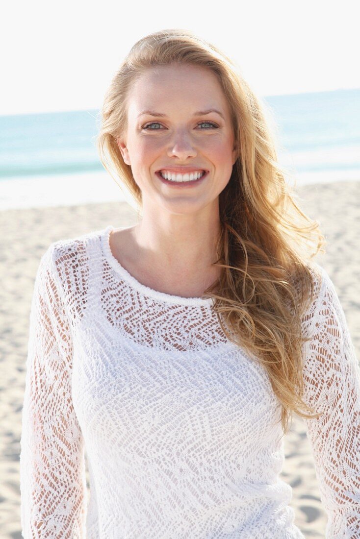 A young blonde woman on a beach wearing a white top and a transparent knitted jumper