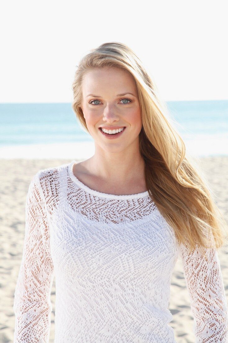 A young blonde woman on a beach wearing a white top and a transparent knitted jumper