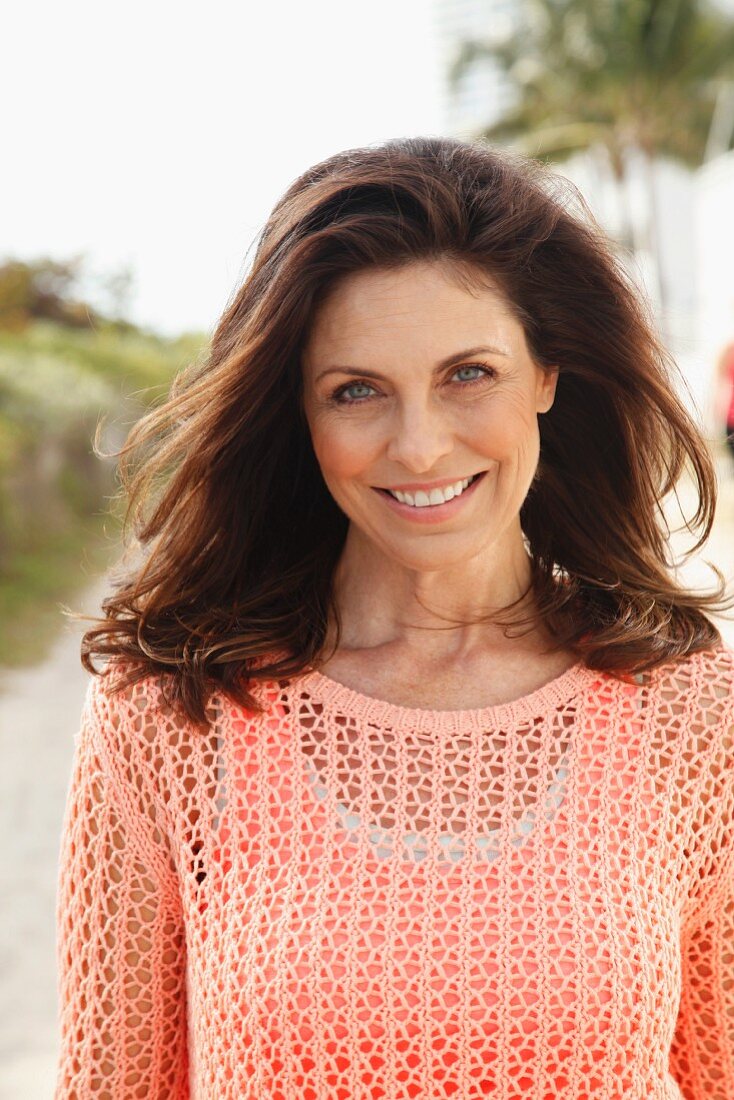 A brunette woman on a beach wearing a salmon-coloured top and an openwork jumper