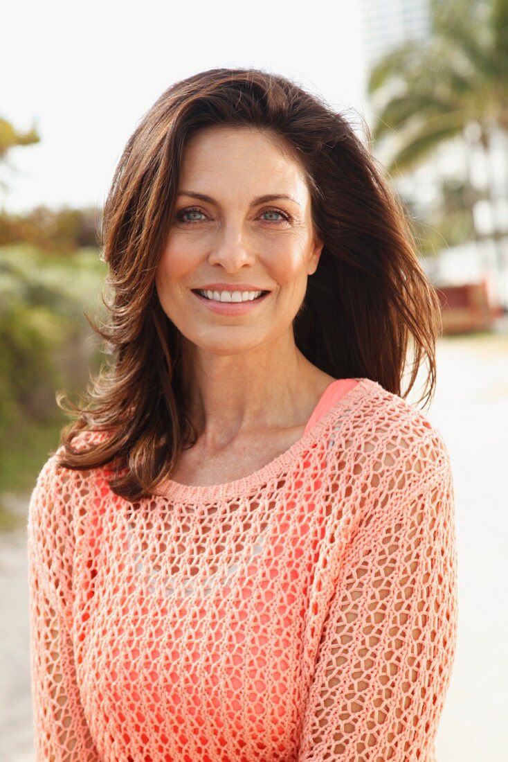 A brunette woman on a beach wearing a salmon-coloured top and an openwork jumper