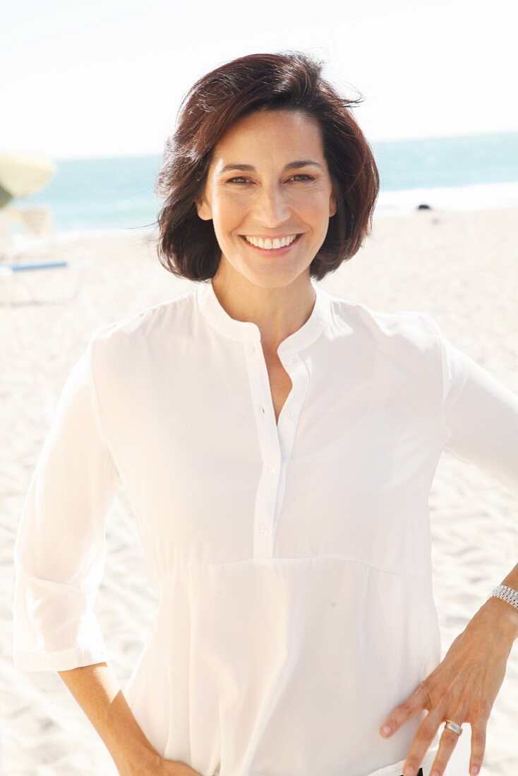 A brunette woman on a beach wearing a thin white blouse