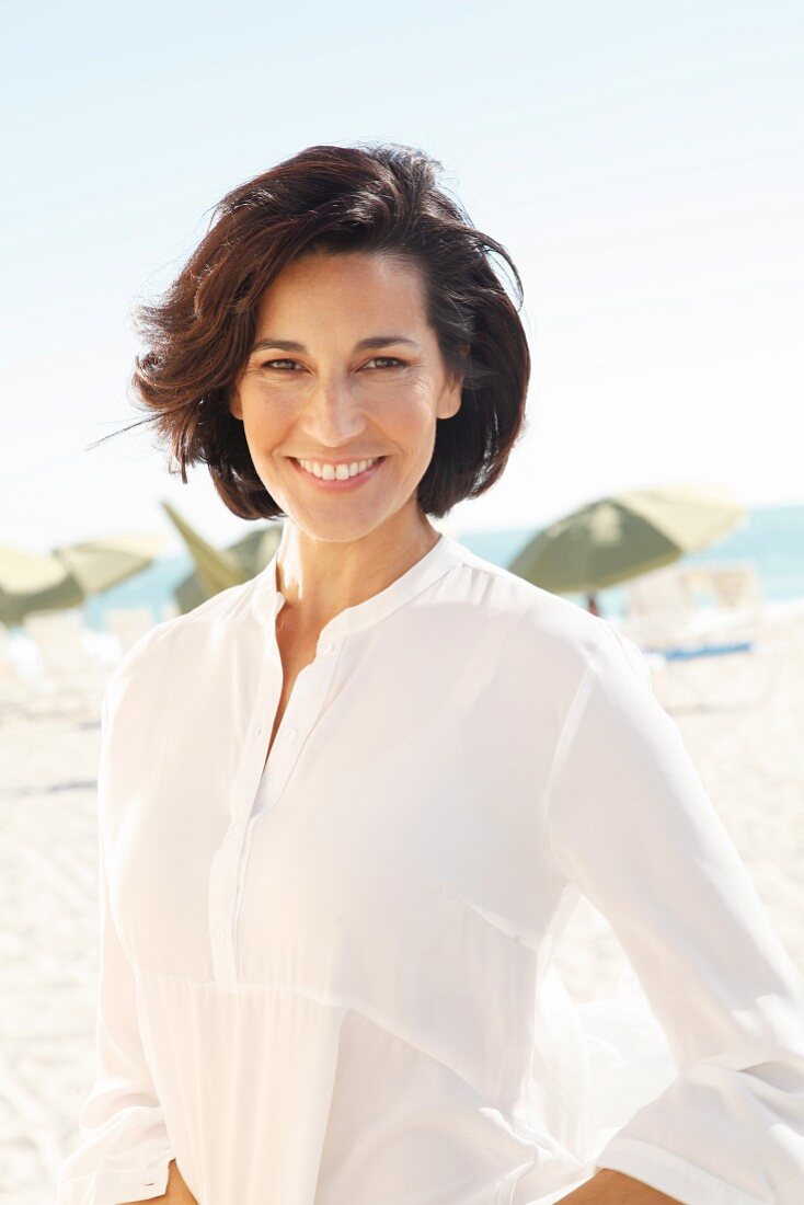 A brunette woman on a beach wearing a thin white blouse