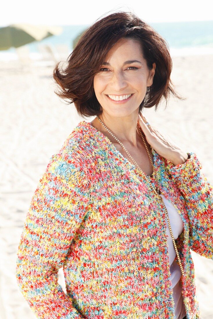 A brunette woman on a beach wearing a white top and colourful cardigan