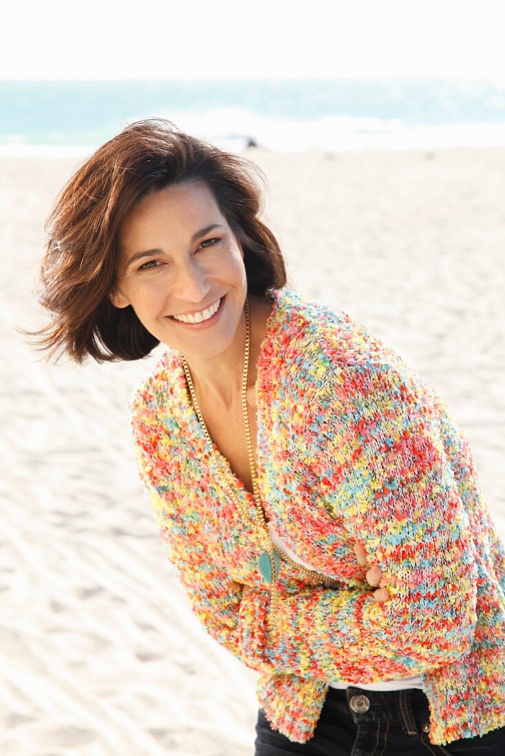 A brunette woman on a beach with her arms folded wearing a white top and a colourful cardigan