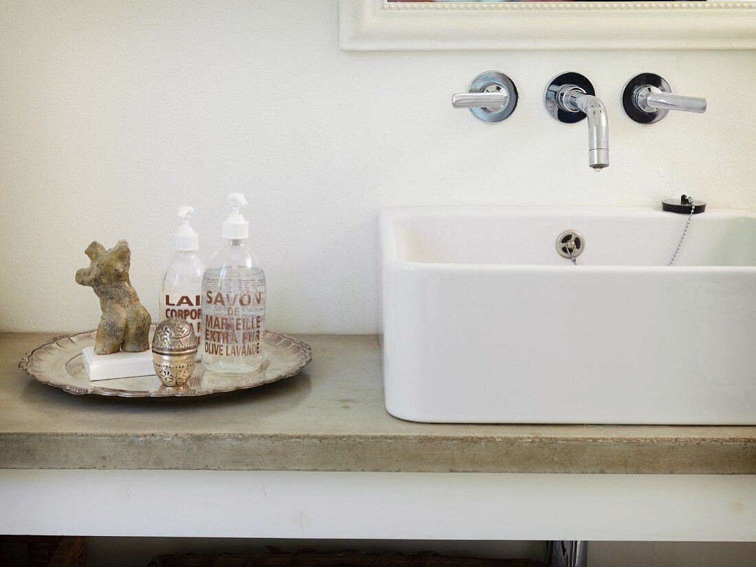 White countertop sink on stone washstand counter next to contemporary soap dispensers and miniature stone torso on vintage-style silver plate