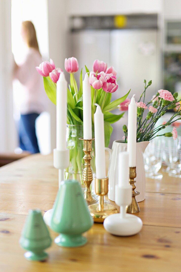 Green salt shakers, white candles in brass and ceramic candlesticks and vases of spring flowers on wooden table